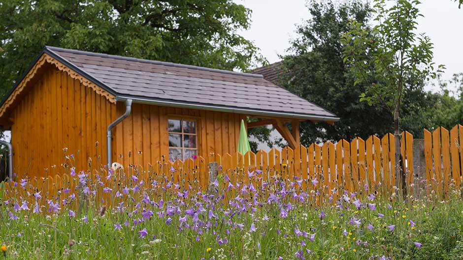 Holzgartenhaus mit Satteldach und Dachrinne, umgeben von Blumenwiese und Holzzaun im Garten