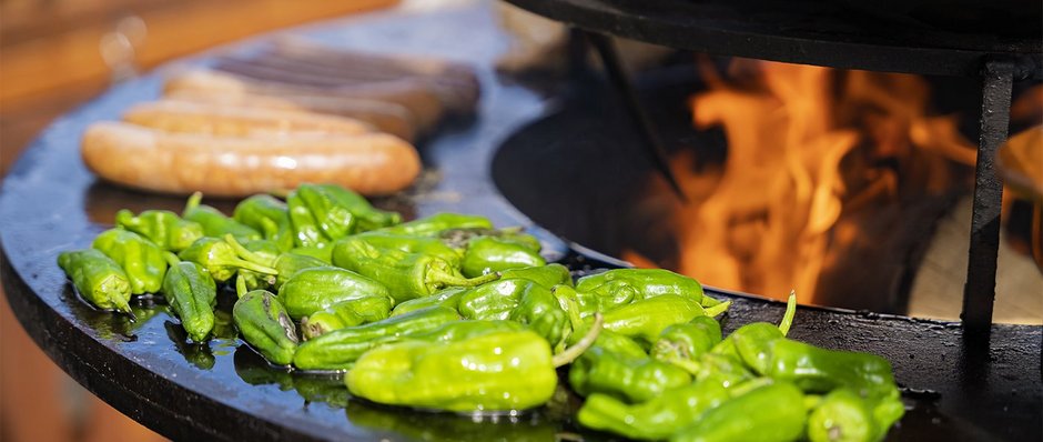 Grüne Paprika und Bratwürste auf Plancha-Grill über offenem Feuer