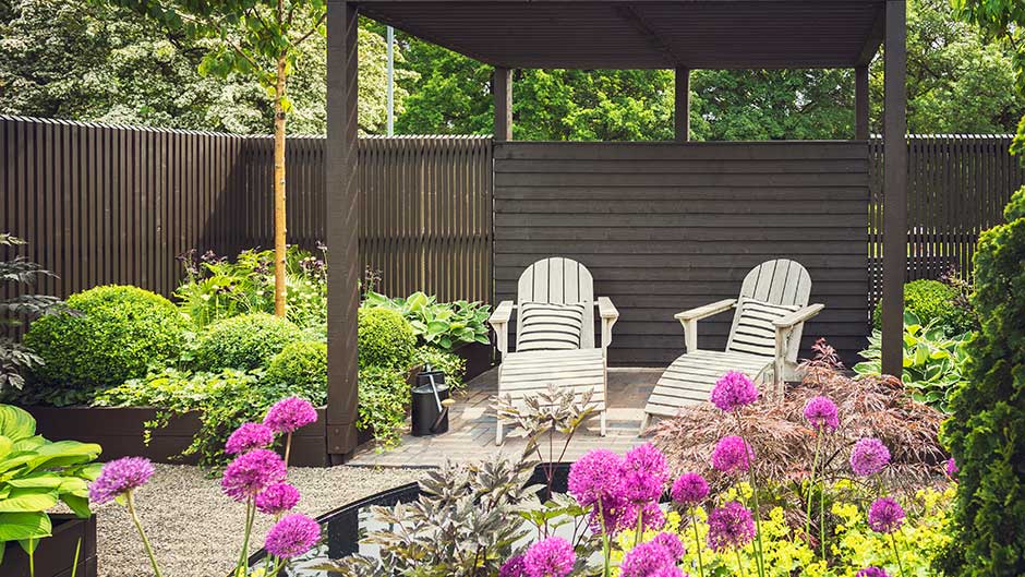 Überdachte Holzterrasse mit zwei Gartenstühlen, umgeben von blühenden Pflanzen