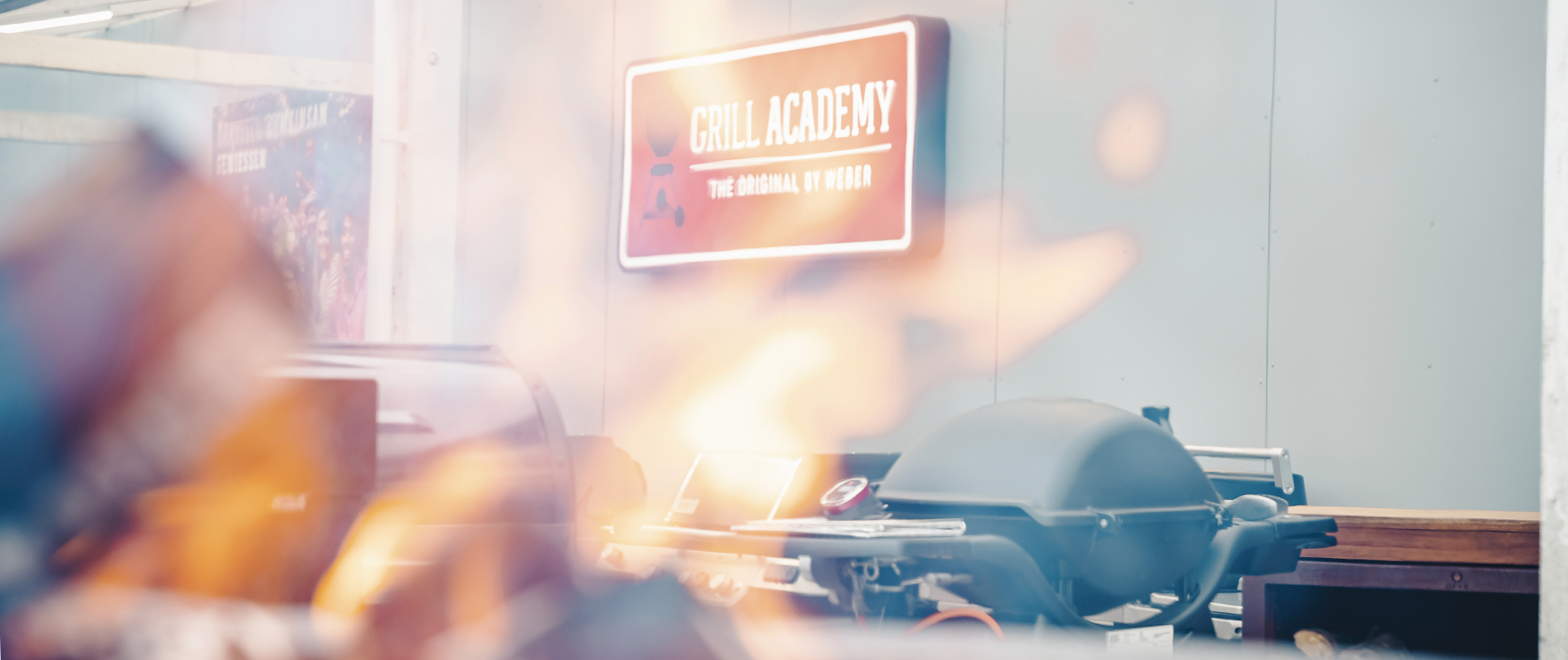 Gasgrill mit Flammen im Vordergrund vor einem Schild mit der Aufschrift ‚Grill Academy‘