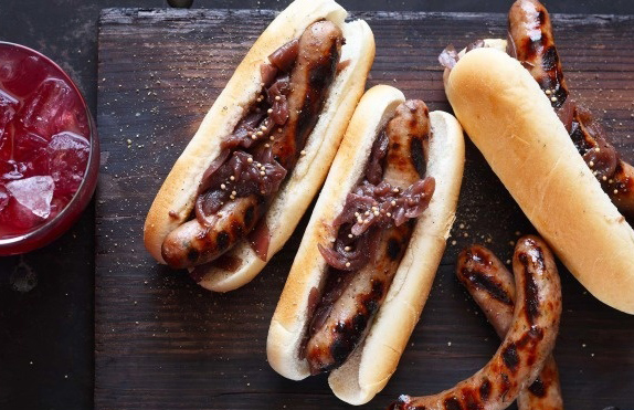 Drei Hotdogs mit gegrillter Bratwurst und Zwiebeln auf einem Holzbrett, daneben ein Glas mit Eisgetränk