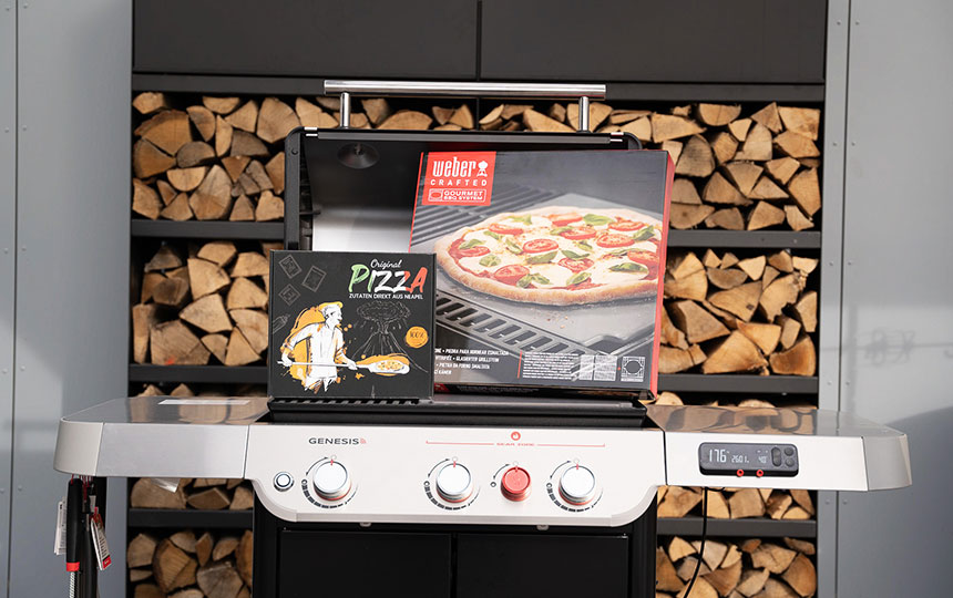 Gasgrill mit Pizzastein und Pizza-Zubehör vor gestapeltem Kaminholz