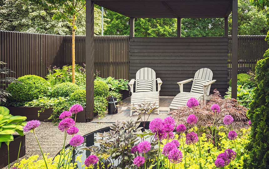 Überdachte Holzterrasse mit zwei Gartenstühlen, umgeben von blühenden Pflanzen